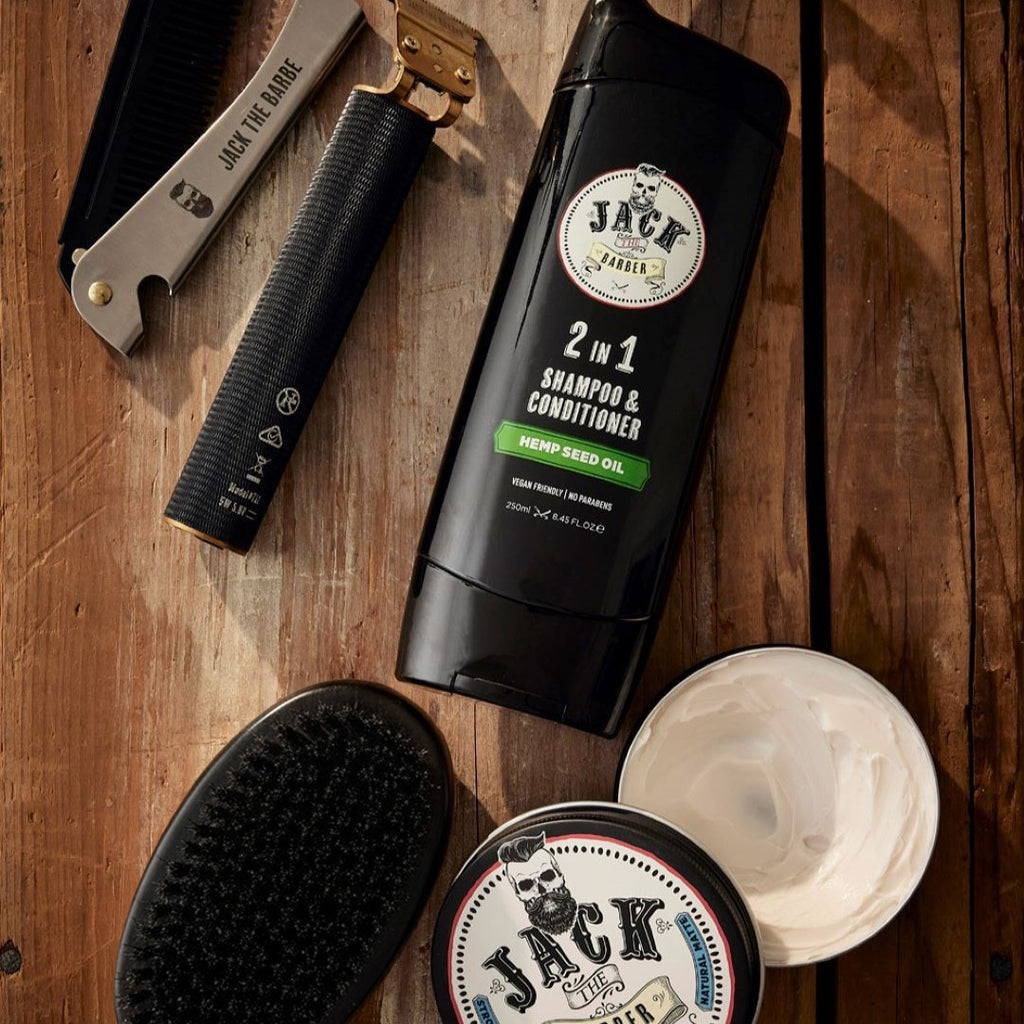 Jack Black grooming products including a 2-in-1 shampoo & conditioner bottle, brush, and jar on a wooden surface.