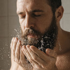 A close-up of a man with a full beard washing his face with water, cupping water in his hands in a bathroom setting.