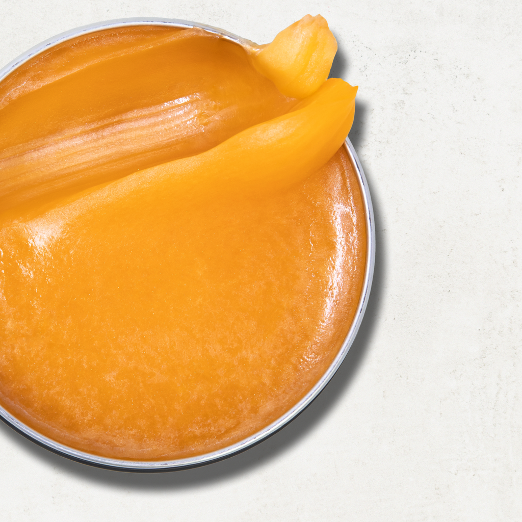 A close-up of a round metal tin filled with a smooth, glossy orange-yellow pomade fill, a thick swirled portion lifted along the rim against a light textured background.