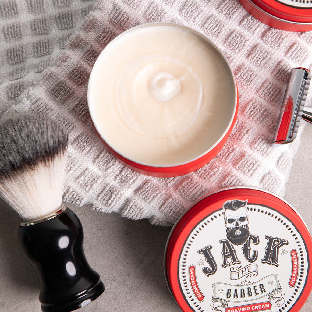 Shaving cream in a red container with a brush and towel on a gray surface, featuring 'Jack the Barber' branding.