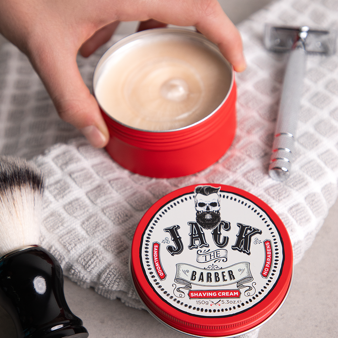 Red container of Jack the Barber shaving cream with a brush and razor on a towel.