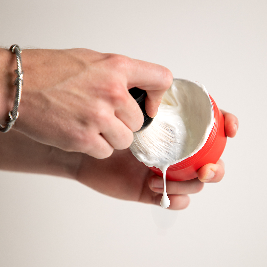 Hand scooping white cream from a red container on a light background